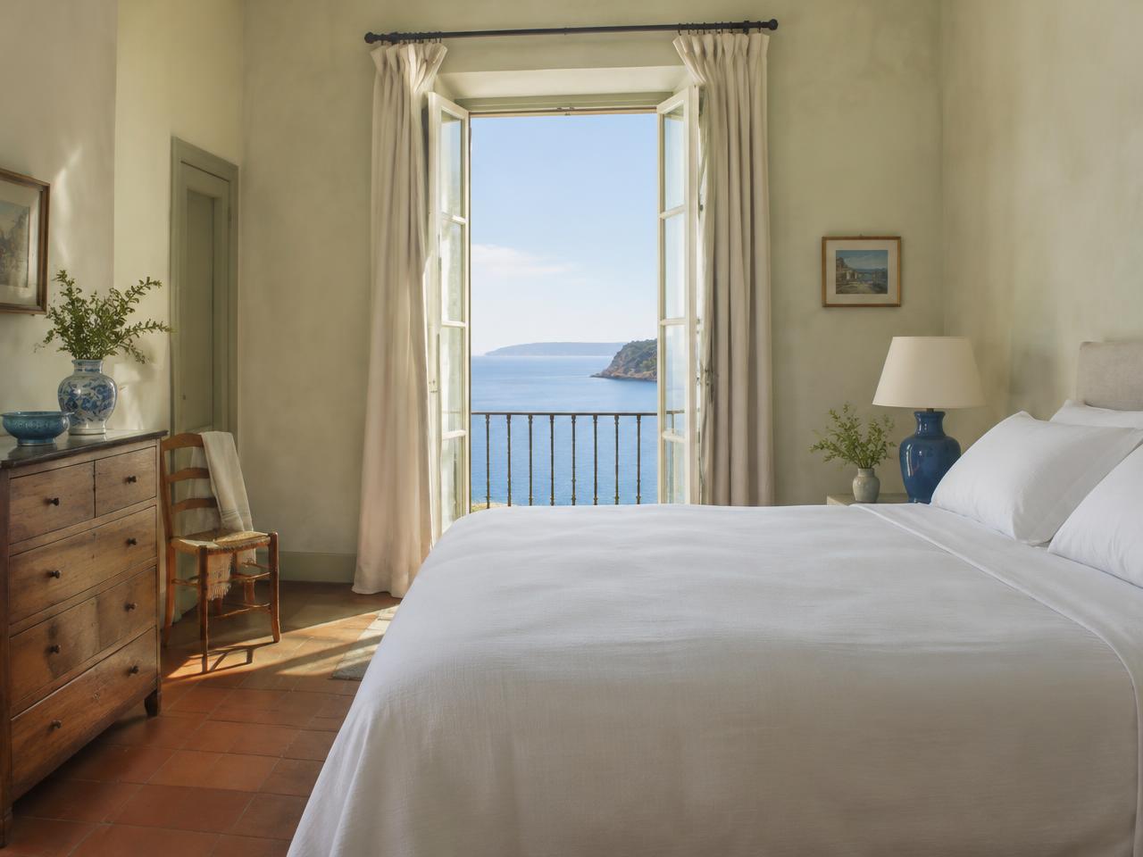 Camera con letto in lino bianco, ceramiche blu e porta-finestra aperta sul mare